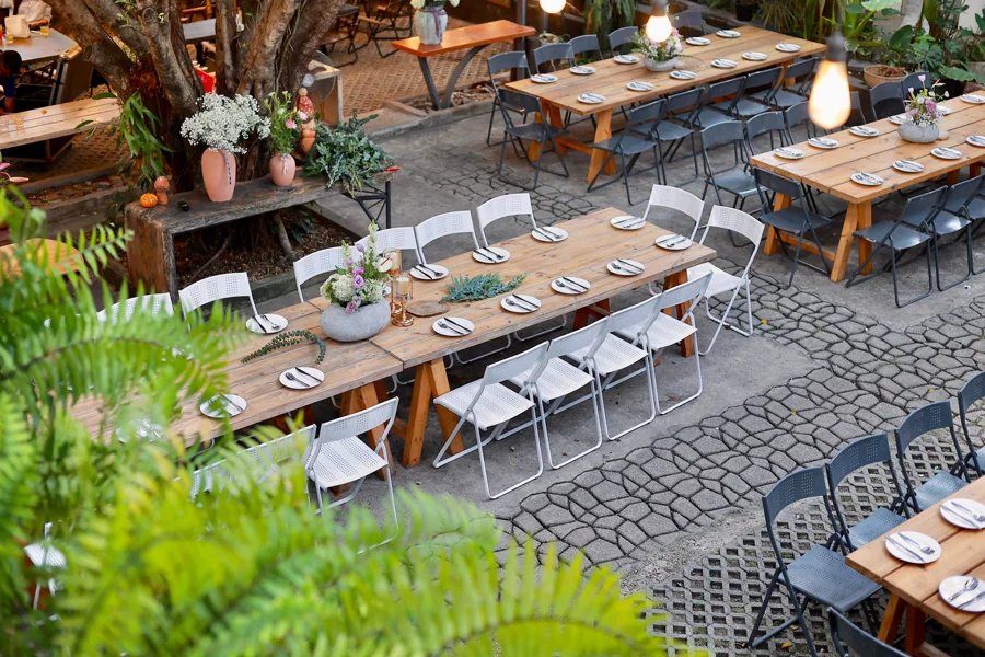Wooden tables and chairs set for an outdoor gathering, surrounded by greenery.