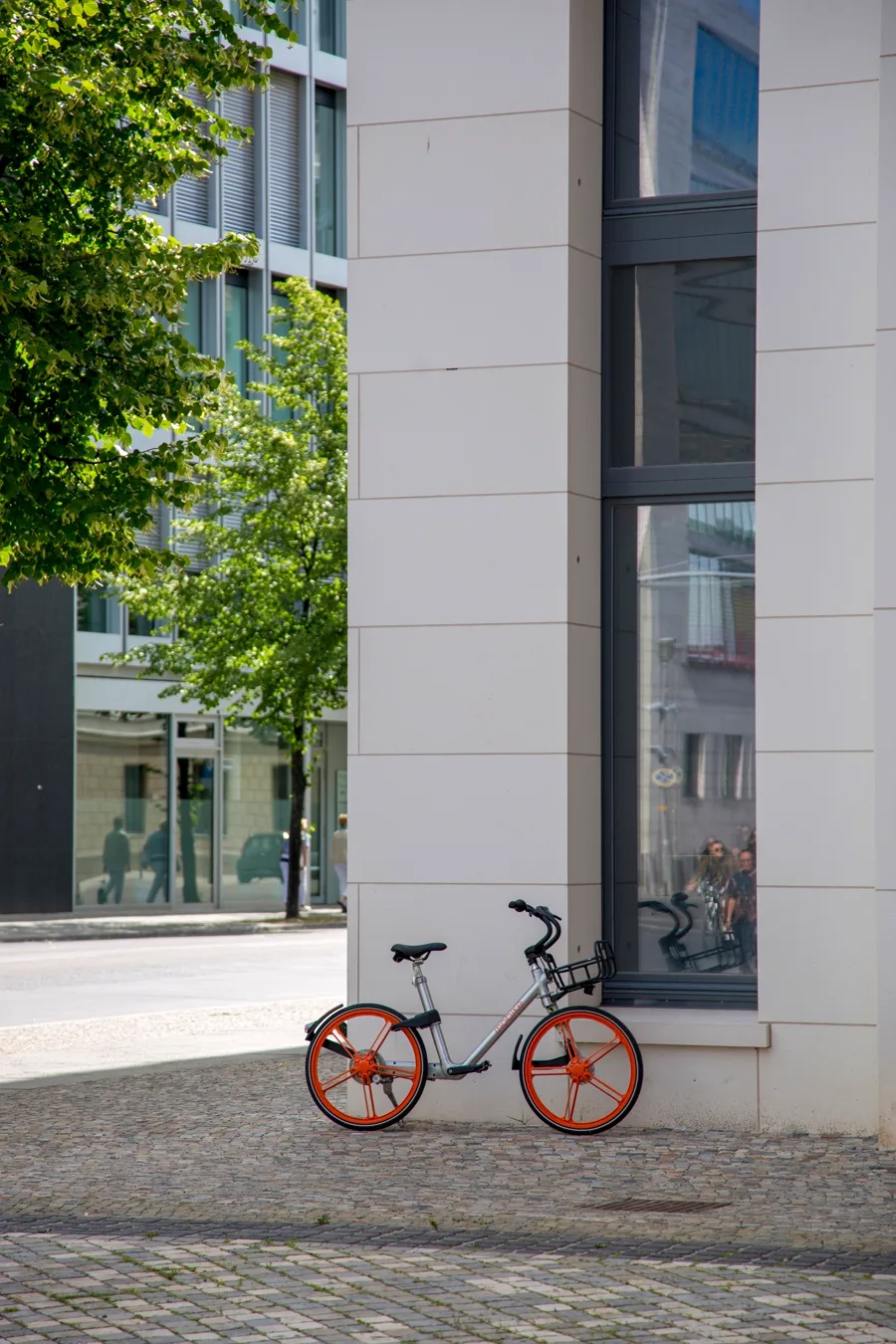 A bicycle with orange wheels stands beside a modern stone and glass building on a cobbled city street.