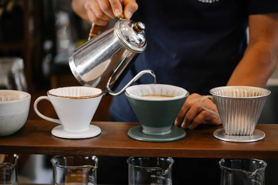 A barista pours from a metal kettle into ceramic coffee drippers on a wooden stand.