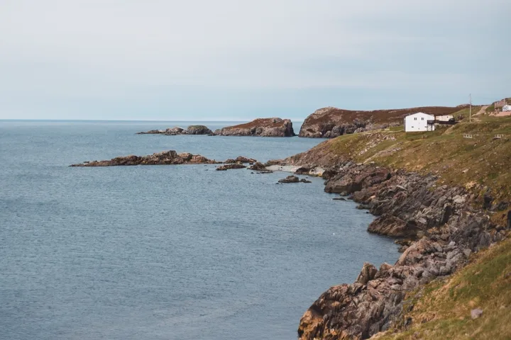Rocky shoreline and a distant white house beside a quiet sea under a cloudy sky.